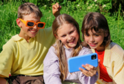 Three smiling children sitting together outside on the grass while taking a selfie on a smartphone.